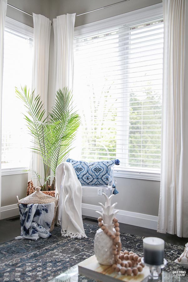 Living room with white drapes, ghost chair, shibori pillow, palm tree, lidded basket, blue area rug.