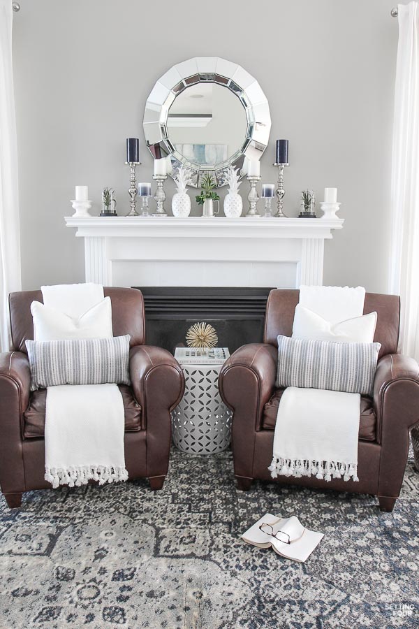 Living room with blue and gray vintage area rug, Pottery Barn leather club chairs, tasseled throws, garden stool, painted fireplace mantel, faceted mirror, mantel decor ideas.