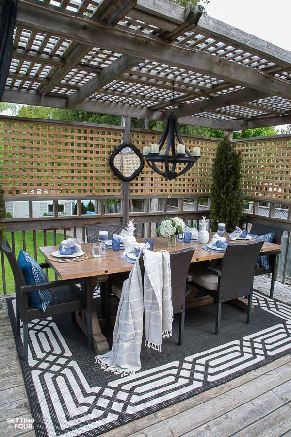 Outdoor gazebo idea for summer. DIY wood Dining table, outdoor chandelier, outdoor mirror, outdoor area rug, wicker stackable chairs, blue and white table setting, planters. 