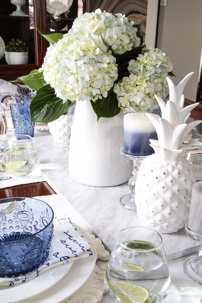 Blue and white napkins, blue bowls, white ceramic pineapple decor, blue hydrangea centerpiece layered together for a summer tabletop idea.