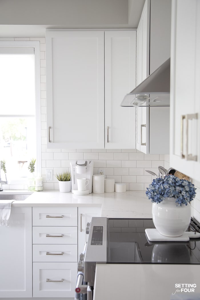 WHite keurig coffeemaker and white kitchen canisters on caesarstone countertops. White kitchen cabinets. Blue hydrangeas. White vase. Kitchenaid electric range with flat ceramic cooktop