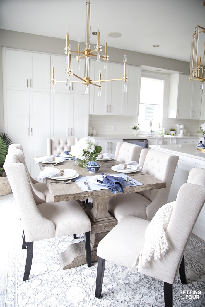 Table settings with a blue and white color palette. Farmhouse table with ivory dining chairs and gold and crystal chandelier.