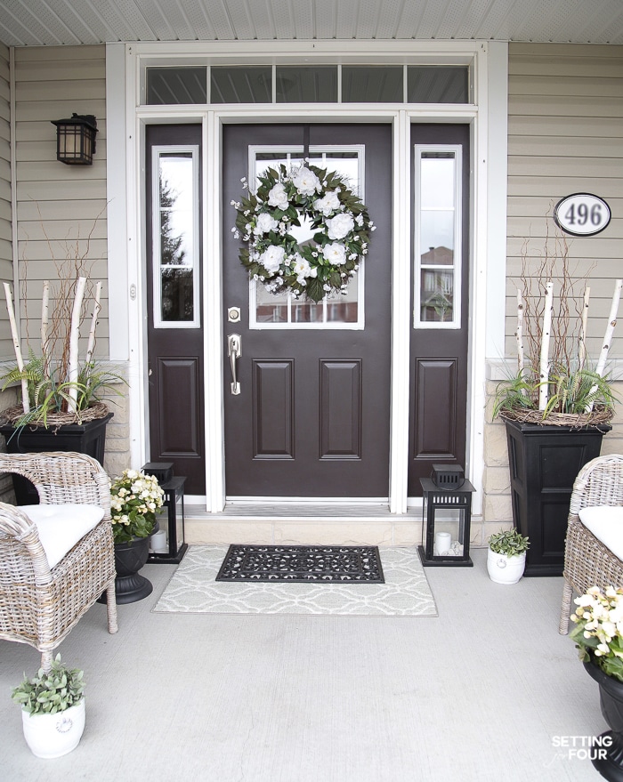 Front porch decorated for summer with a peony wreath hung on the front door. Planters decorated with birch branches and ferns. Wicker chair seating.