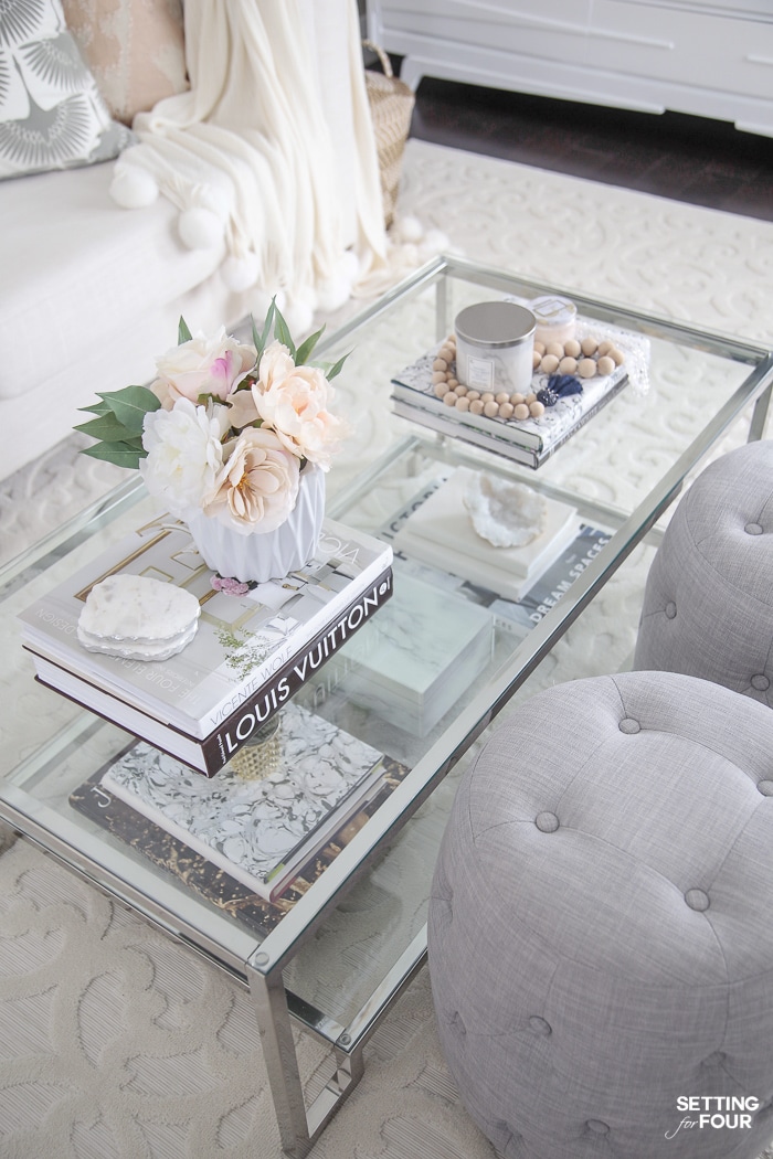 Tiered glass coffee table decorated with stacks of books, candles, coasters and vase of flowers.