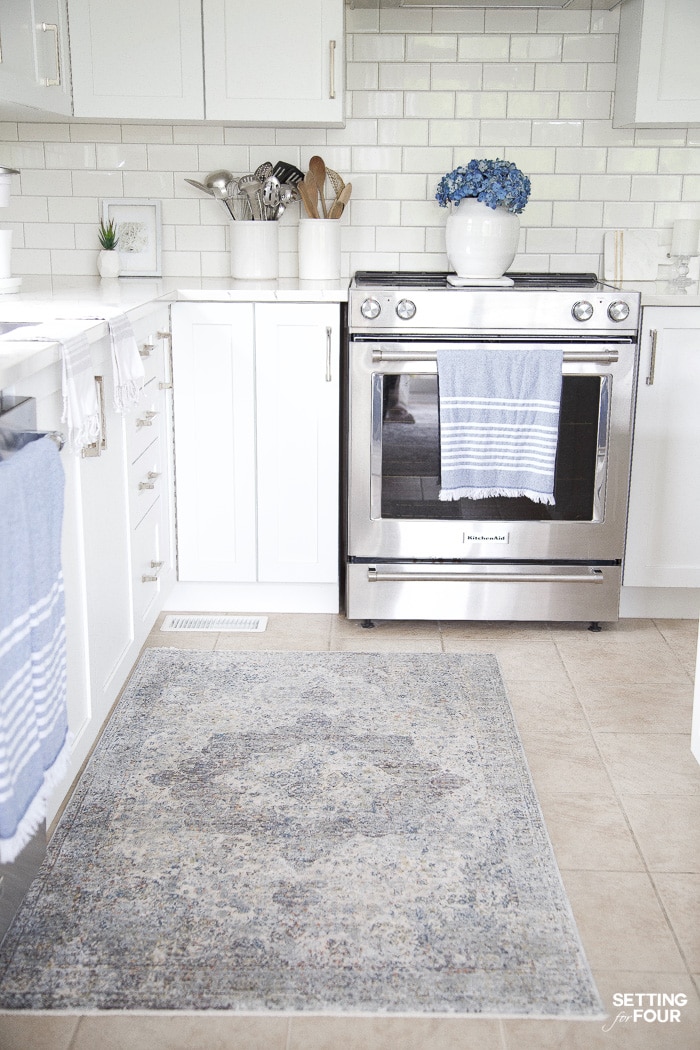 Kitchen with white cabinets. White subway tile with gray grout. Blue and white medallian kitchen runner rug. Blue tea towels. Blue hydrangeas in a white vase.