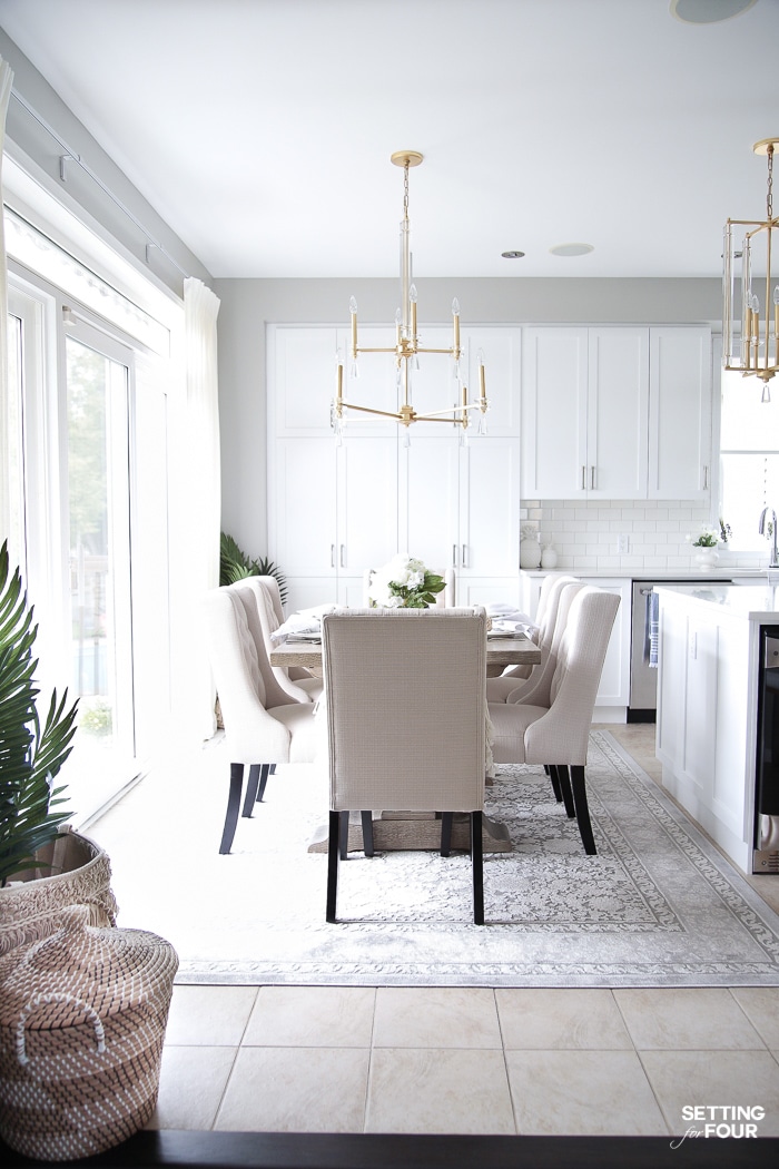 Dining area decorated for summer with blue and white tablescape. Gold and crystal chandelier hanging over gray wood dining table. Gray rug and upholstered dining chairs.