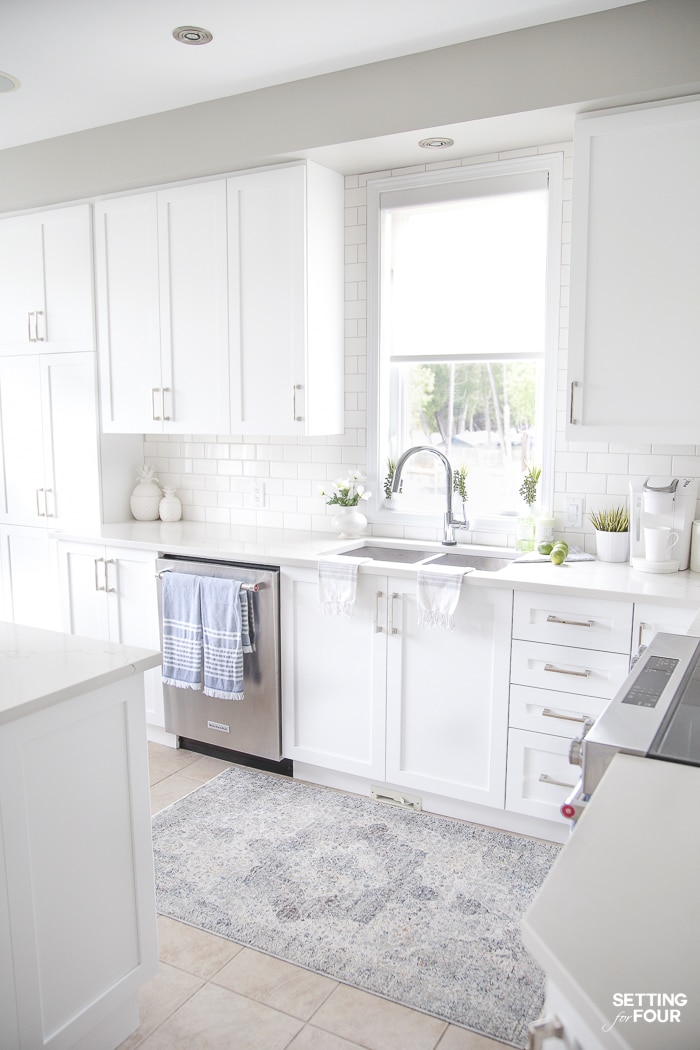 Blue and white striped tea towels hung on a dishwasher and oven in a white kitchen. Blue rug in front of sink. Pineapple canisters and summer kitchen decor.