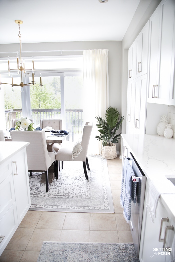 Open concept kitchen and dining area. Restoration Hardware wood dining table with ivory dining chairs, crystal and brass chandelier and palm trees in baskets.