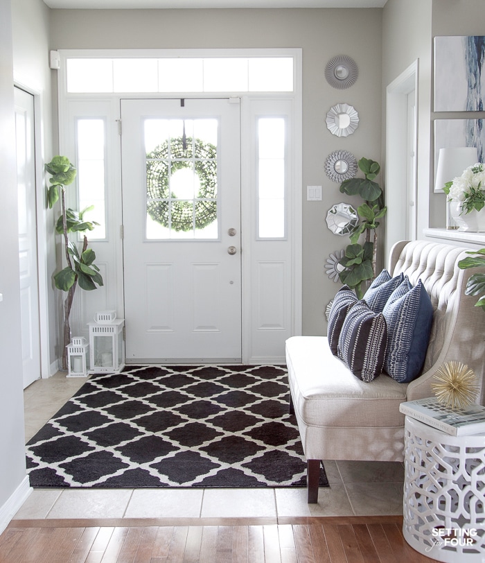 A birght entryway with area rug, plants, setee for seating and blue pillows for color and pattern