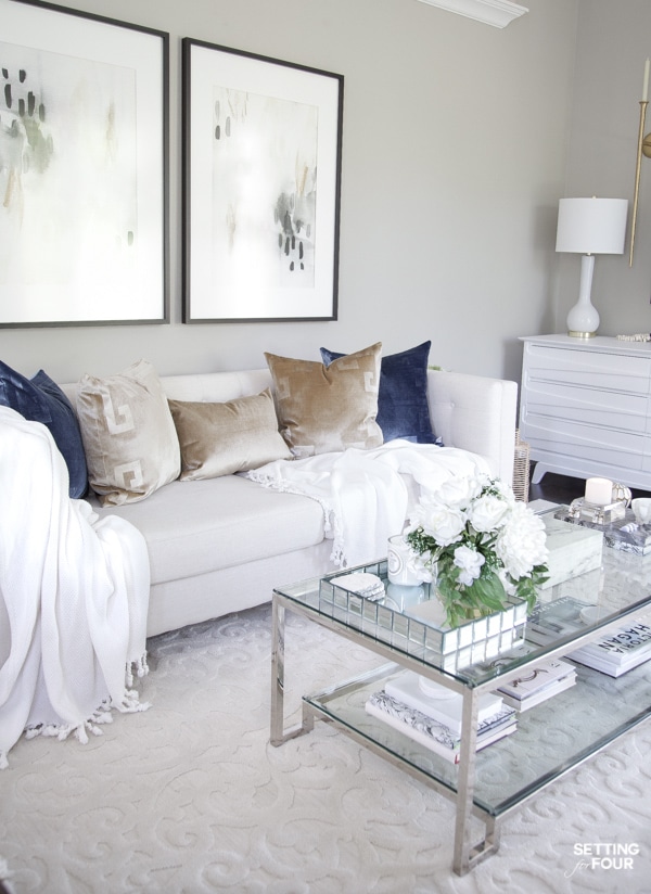 Summer living room decorating ideas with blue and gold pillows, white throw blankets, abstract wall art, coffee table decor. Sherwin Williams Mindful Gray paint color on walls