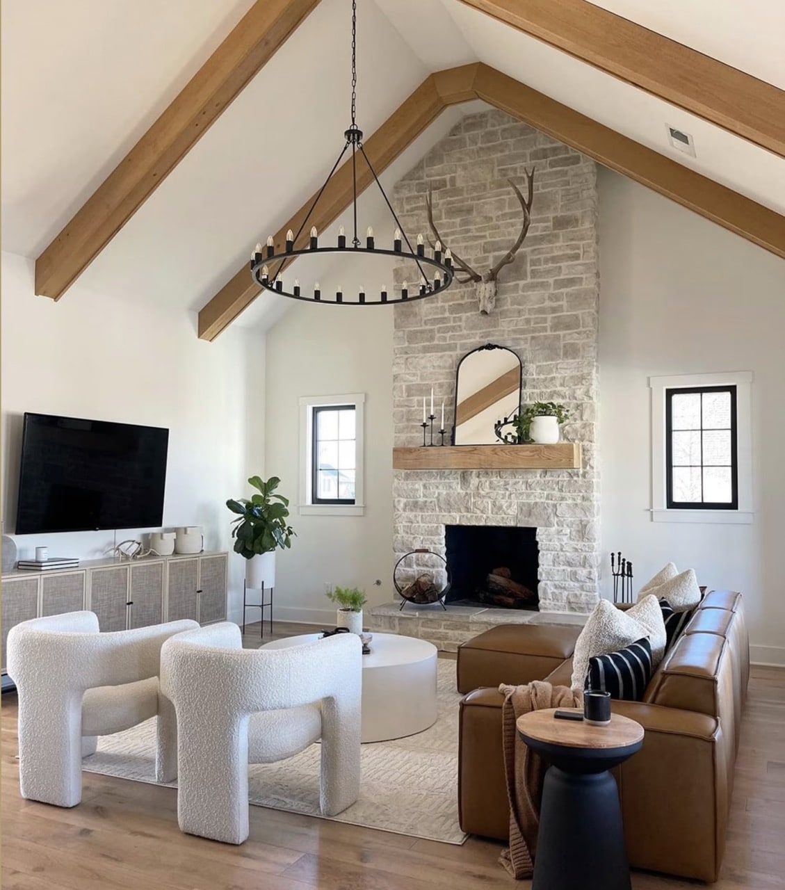 Sectional Sofa With 2 Chairs And TV Unit - Living Room Furniture Placement Idea , vaulted ceiling with wood beams, tall natural stone fireplace with wood mantel and mirror above mantle, windows on each side of fireplace, rattan tv unit cabinets, boucle chairs, leather sectional chaise, cozy, earthy color palette, warm color scheme, black round chandelier, transitional, organic modern, farmhouse, traditional, minimalist home inspiration, new home build