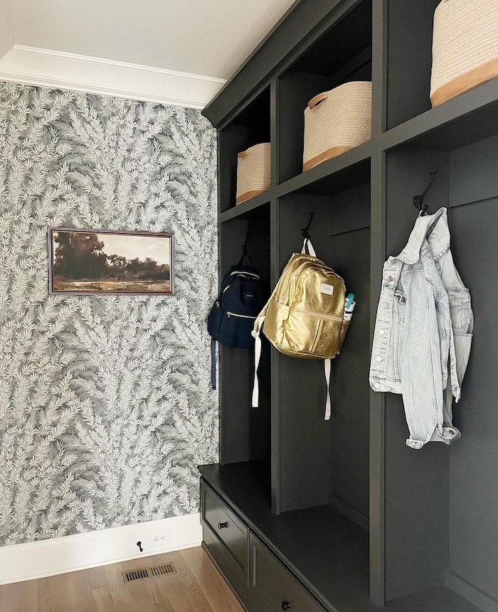 Beautiful Mudroom Storage Ideas For Backpacks and Coats. A stylish mudroom with built in dark cabinets featuring hooks for backpack storage, baskets and coats. Neutral wallpaper and landscape art add elegance.
