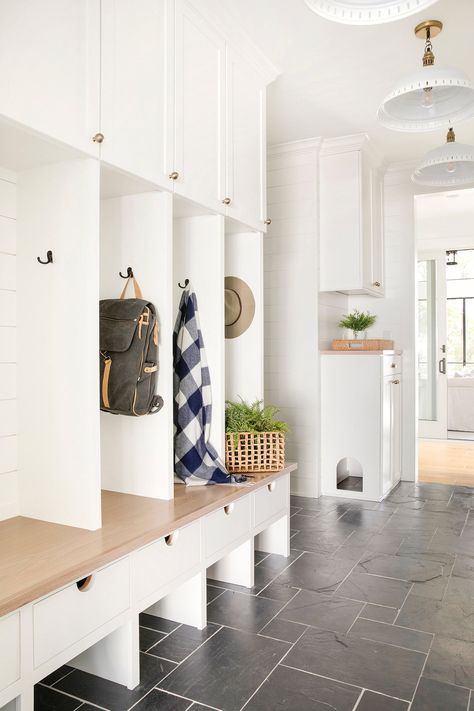 Stylish mudroom organization ideas with white built in cabinets, black hooks holding a backpack, hat, scarf, wicker baskets, slate tile floor, and bright lighting.
