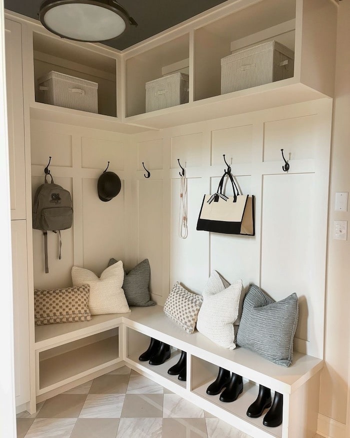 Cozy white mudroom storage ideas with built-in bench, plush cushions, and boots neatly arranged below. Hooks on board and batten wall hold backpacks, purse, hats and dog leash while upper shelves store lidded boxes for extra storage.