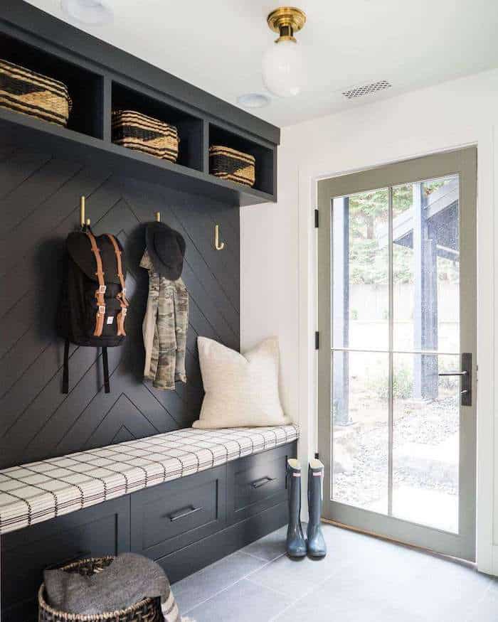 Mudroom storage ideas for backpacks, coats, and shoes. Black built in cabinet with bench and cushion. Wall hooks hang backpacks and jackets. Shelves have woven baskets for additional storage. 
