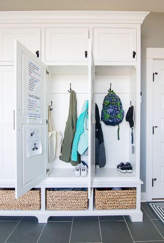 Beautiful mudroom built in lockers idea. White clocker cabinets for backpack storage, coat shoe and hat storage. Baskets in open cubbies store shoes underneath and upper cabinets with doors store gloves and hats for a calm, tidy look. A family schedule and calendar organize kids activities.