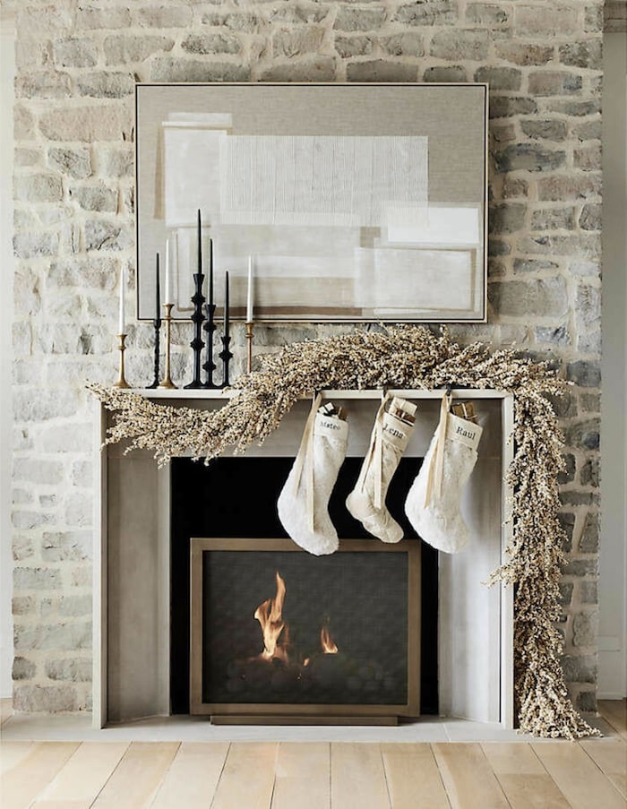 Elegant, neutral Christmas mantel decor ideas in a cozy living room with white berry garland and white Christmas stockings. Natural stone fireplace wall and beautiful candles on the holiday mantle. 