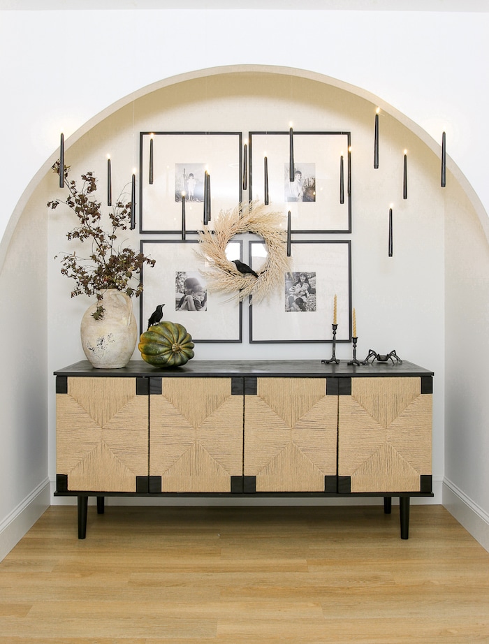 Minimalist decor with a woven-texture sideboard, arch framing. Wall displays photos, wreath, and black candles for an elegant, cozy atmosphere. Modern Halloween decor ideas for the entryway or hallway.