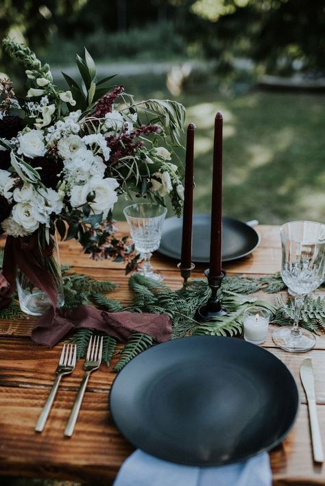 Elegant outdoor table setting with a rustic wooden table, dark plates, and black candles. Features lush floral centerpiece, greenery, and wine glasses. floral halloween centerpiece.