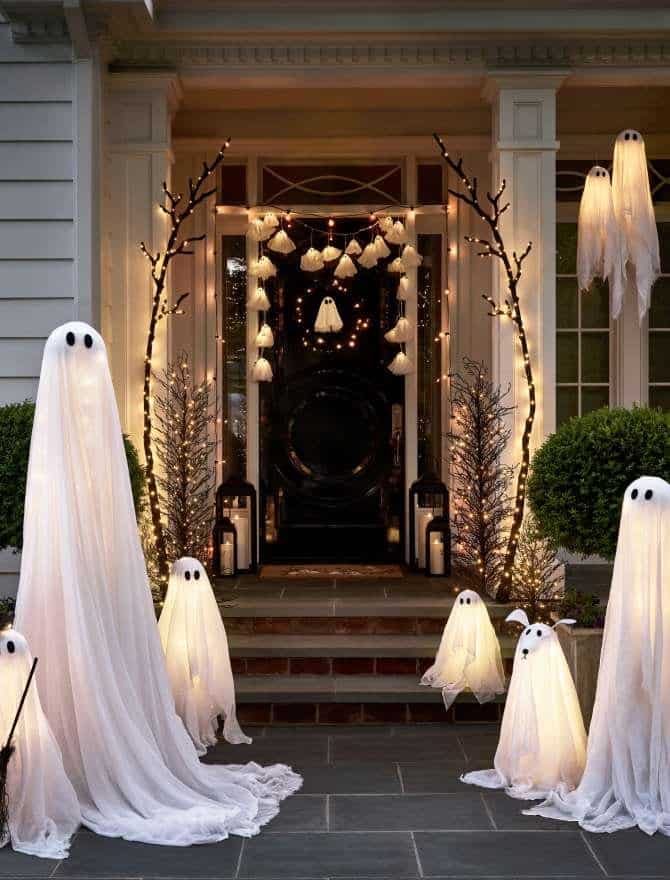 Front porch decorated for Halloween with lit ghost figures and lanterns. Ghosts are surrounded by lit branches, creating a spooky, festive atmosphere. lit black trees.