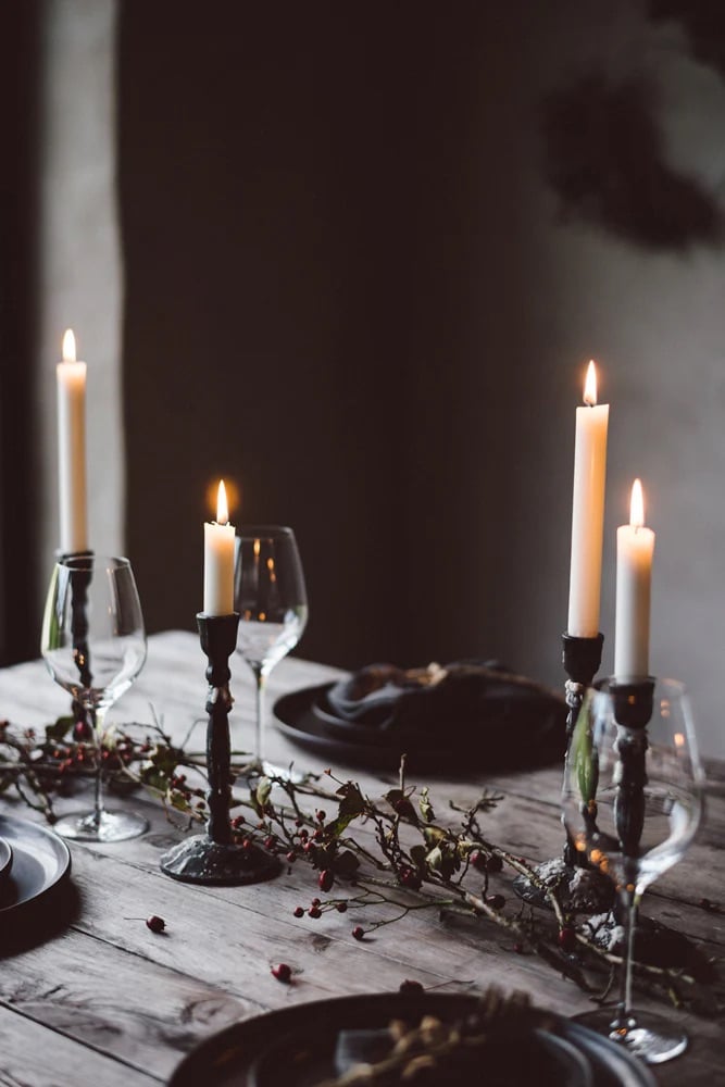 A rustic wooden table is elegantly set with tall black candle holders and lit candles, surrounded by dark plates and wine glasses. A garland of red berries adds a cozy, romantic touch. Elegant Halloween Tablescape Decor Ideas