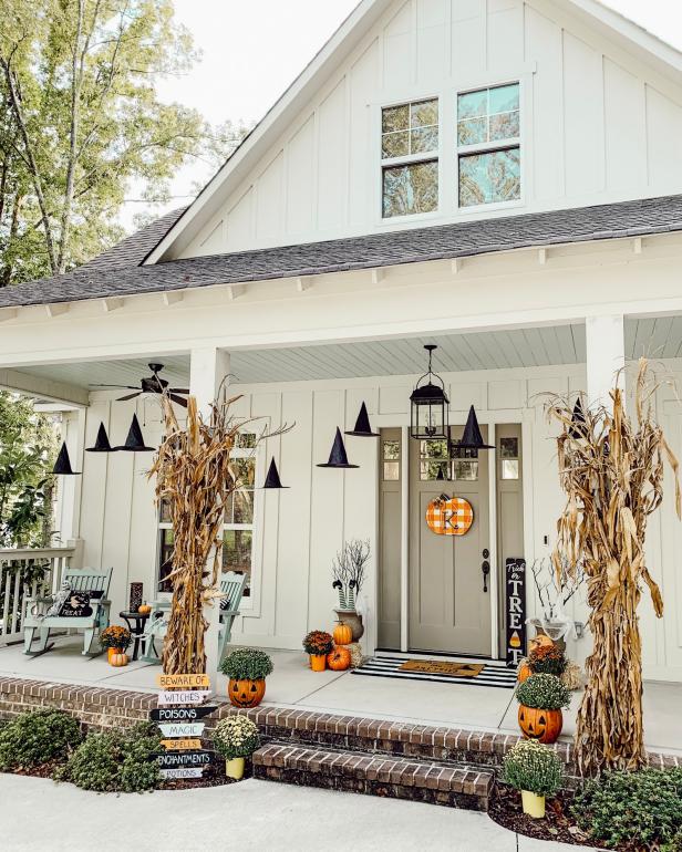 Charming Halloween porch with a white house backdrop. Decor includes hanging witch hats, pumpkins, cornstalks, playful signs, and autumn plants. Cozy and festive.