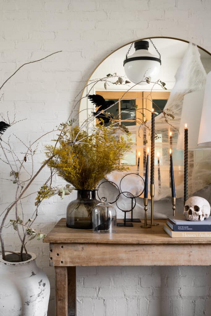 A rustic wooden table with fall flowers, black candles, and a skull under a round mirror. A black bird, branches in a vase and cobwebs add a spooky, autumnal vibe.