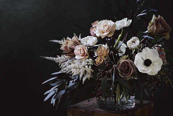 A lush floral arrangement in a glass vase features cream roses, white anemones, and eucalyptus leaves against a dark background, conveying elegance and serenity.
