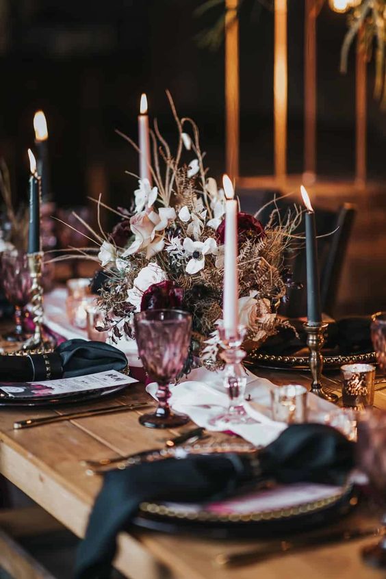Elegantly set dining table with dark candles, ornate floral centerpiece, and purple glassware on a wood dining table. The mood is romantic and sophisticated.