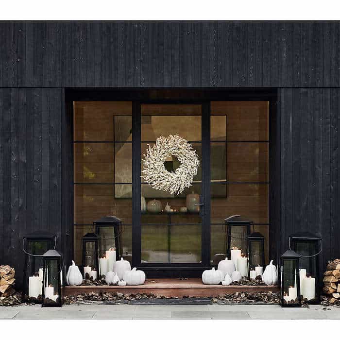 Modern black steel and glass door with a white wreath. Surrounded by white pumpkins and large lanterns with lit candles, creating a cozy autumn atmosphere. Fall porch decor ideas.