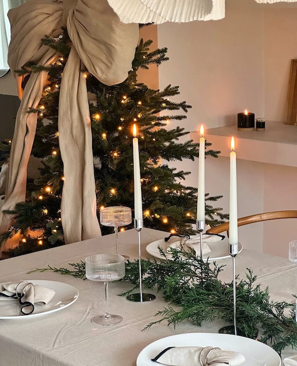 Elegant, Neutral Christmas Dining Table Decorating Ideas. Simple Centerpiece of garland and taper candles. Christmas tree with large bow Christmas 2024 decor trend.