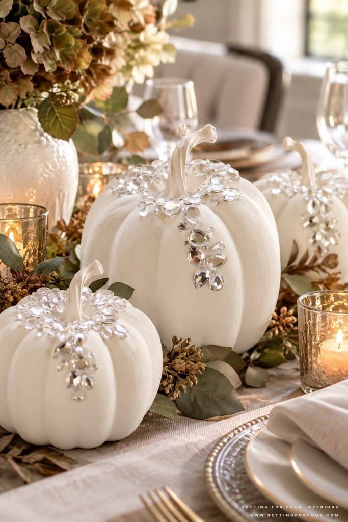 Elegant fall dining table centerpiece featuring three white painted faux pumpkins decorated with cascading clear rhinestones, surrounded by eucalyptus greenery, dried hydrangeas and glowing gold votive candles. Neutral linen table runner, layered white dinnerware and gold accents create a luxurious Thanksgiving tablescape with sophisticated DIY no-carve pumpkin decorations.