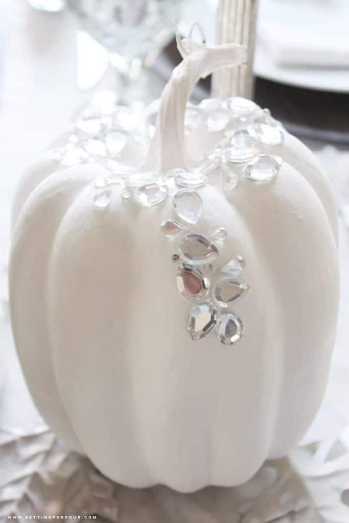 Close-up of an elegant white painted faux pumpkin decorated with sparkling clear rhinestones cascading from the stem down the front. The DIY no-carve pumpkin is displayed on a silver metallic leaf placemat with soft white table linens and crystal glassware in the background, creating a sophisticated fall and Halloween decorating idea.