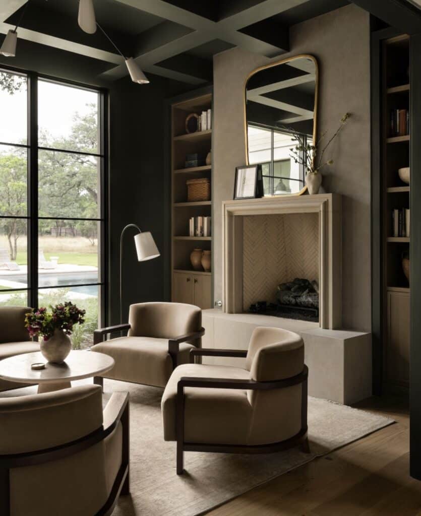 Moody organic modern living room with coffered ceiling, neutral upholstered chairs, round coffee table, built-in shelving, modern fireplace with herringbone detail, large window, and layered textures.