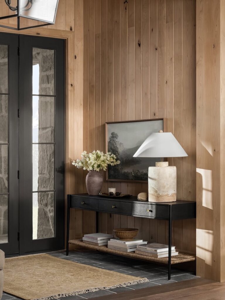 Warm organic modern entryway with vertical wood paneled walls, black framed glass front door, black console table, sculptural stone table lamp, framed artwork, floral arrangement, woven lower shelf, and neutral area rug.