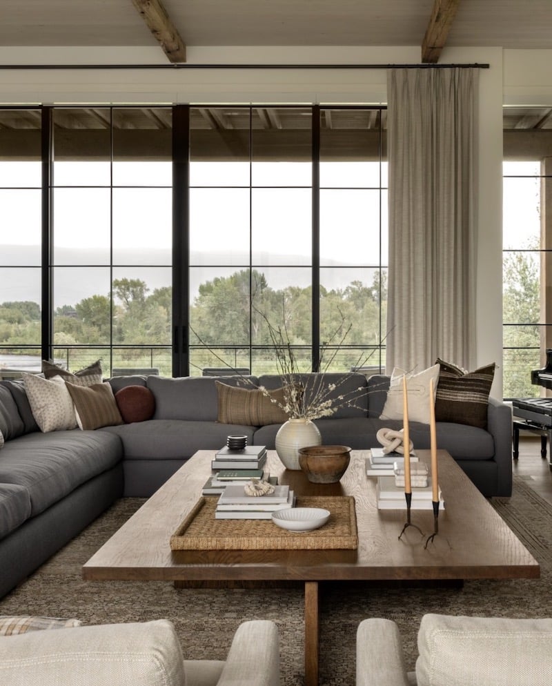 Neutral organic modern great room with oversized gray sectional sofa, large wood coffee table, layered throw pillows, floor-to-ceiling black framed windows, linen drapery, and warm wood accents.