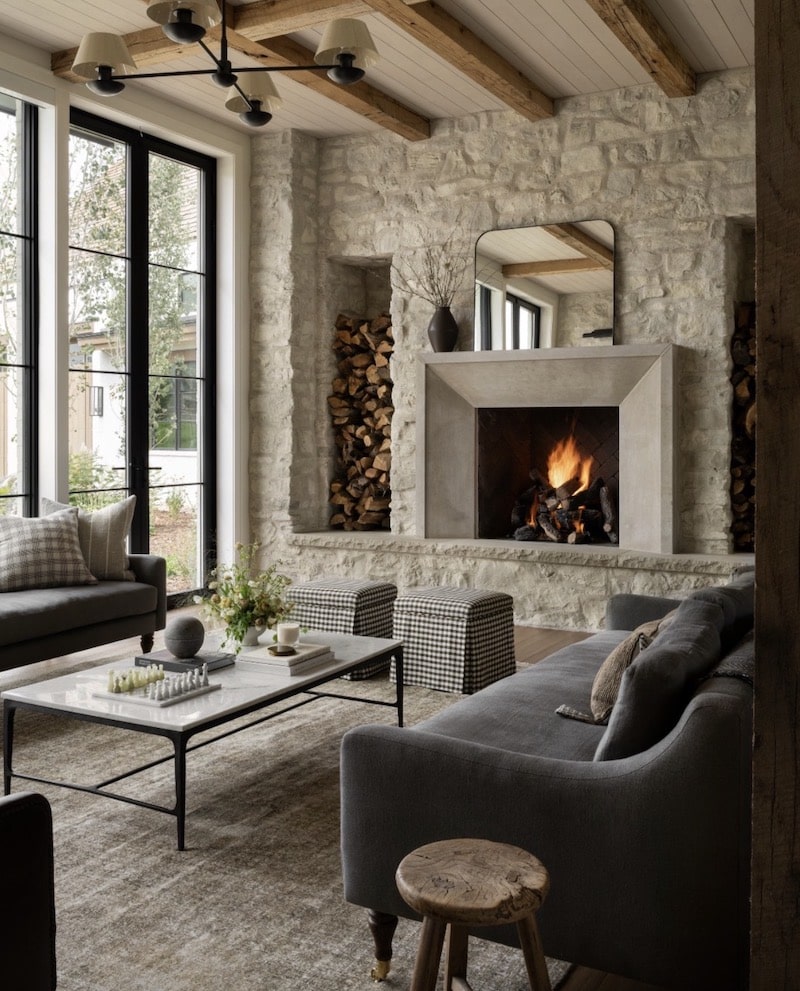 Cozy neutral living room with a large stone fireplace, warm wood ceiling beams, black framed windows, gray upholstered seating, marble coffee table, checkered ottomans, and layered organic modern decor.