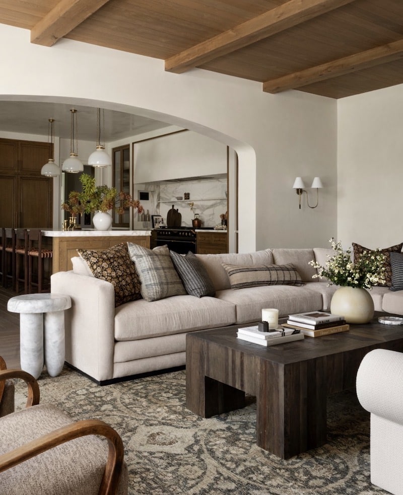 Neutral organic modern living room with warm wood ceiling beams, cream sectional sofa, dark wood coffee table, patterned area rug, layered throw pillows, and an open archway view into a warm-toned kitchen with pendant lights.