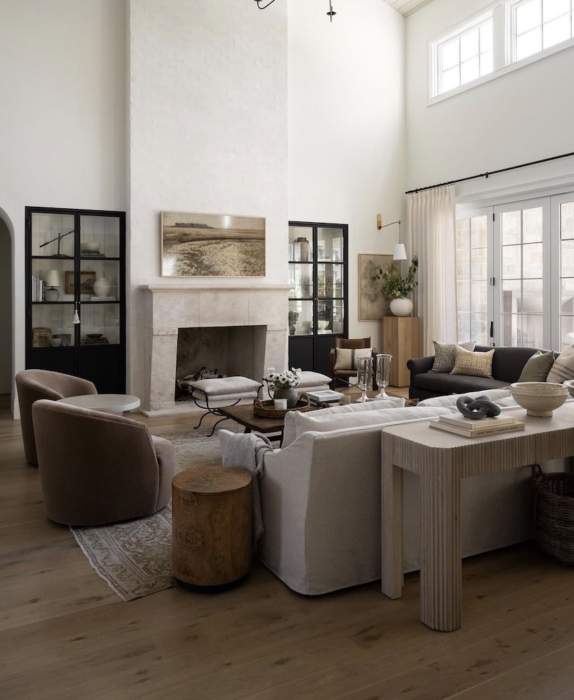 Warm neutral living room with a tall fireplace, white sofa, curved accent chairs, wood floors, and layered decor showing how to arrange living room furniture with a fireplace.