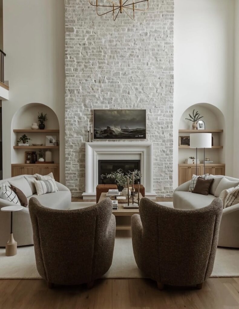 Neutral living room with white sofas, brown accent chairs, wood shelves, and a tall stone fireplace showing a balanced living room furniture arrangement with a natural stone fireplace.