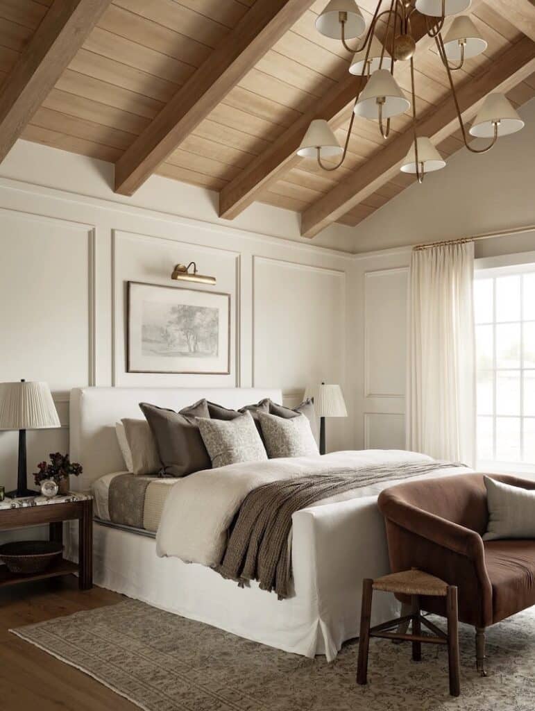 Neutral organic modern bedroom with warm wood ceiling beams, cream upholstered bed, layered neutral bedding, paneled walls, brass chandelier, soft linen curtains, patterned area rug, and warm brown accent chair.