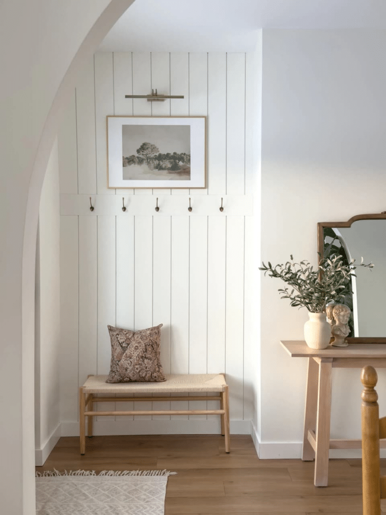 Create an entryway in your home when you don't have one! This small nook entryway idea is beautiful. Add shiplap wall pannelling with wall hooks to a recessed wall to add storage for coats and scarves. A small bench underneath adds a spot to sit and put on shoes. Add wall art with a picture light above the wall hooks to complete the look! Modern heritage, farmhouse, new traditional, organic modern design style, Nancy Meyers style, white walls, bright hallway, modern entry inspiration, 2026 design ideas, neutral color palette, warm earth tones color scheme.