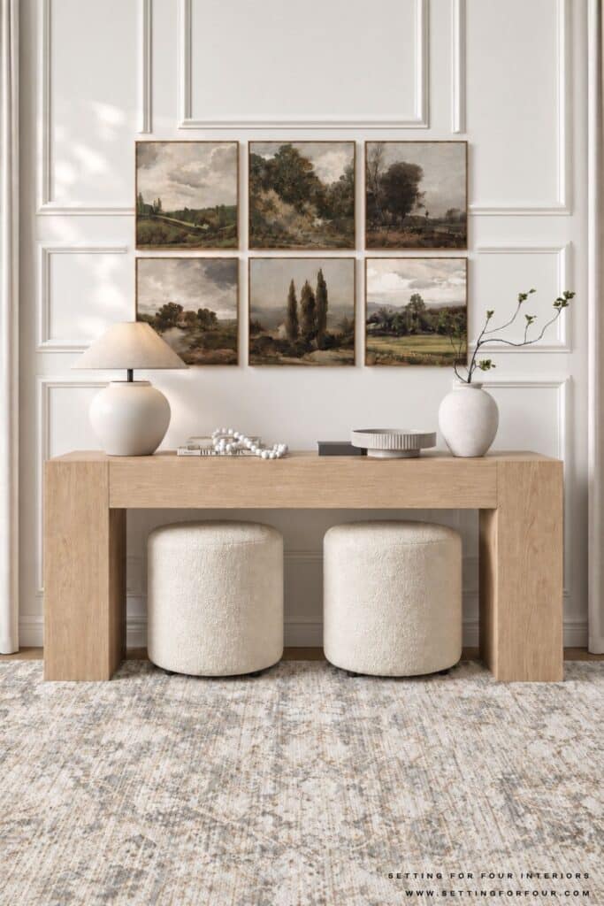 White oak console table styled beneath a six-piece landscape gallery wall with a cream lamp, books, decorative bowl, greenery and two cream ottomans.