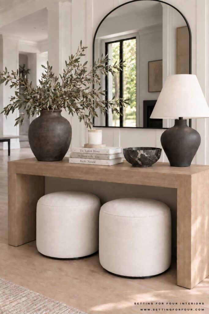 White oak entryway console table styled with an arched black mirror, black lamp, olive branches, books, candle, marble bowl and two cream ottomans.